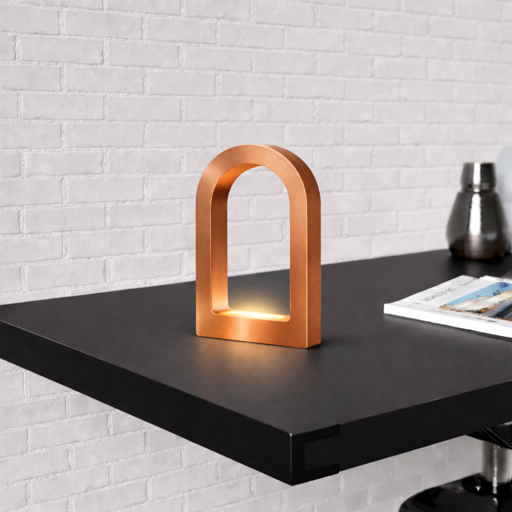 U-shape curved desk lamp with copper finish by WOOM, emitting warm light on a black table against a white brick wall.
