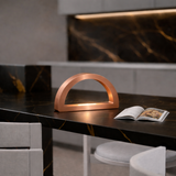 Copper finish arc-curved desk lamp by WOOM, emitting warm light on a black marble counter with golden veins.