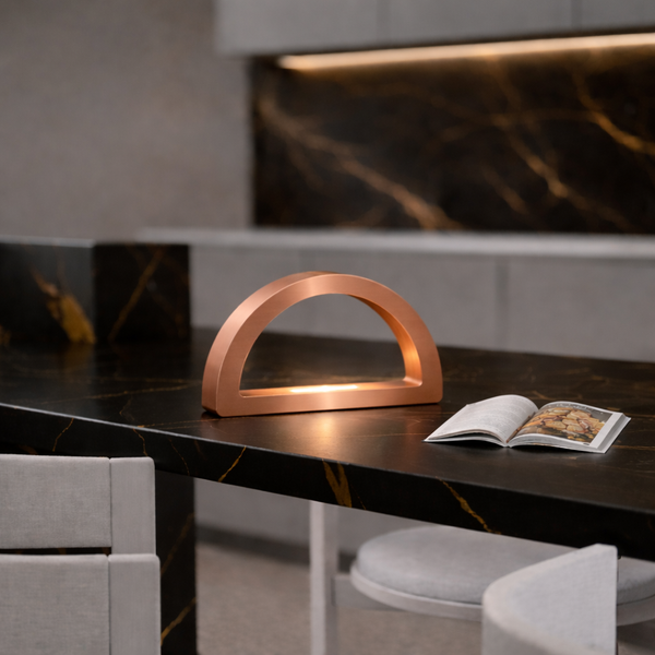 Copper finish arc-curved desk lamp by WOOM, emitting warm light on a black marble counter with golden veins.