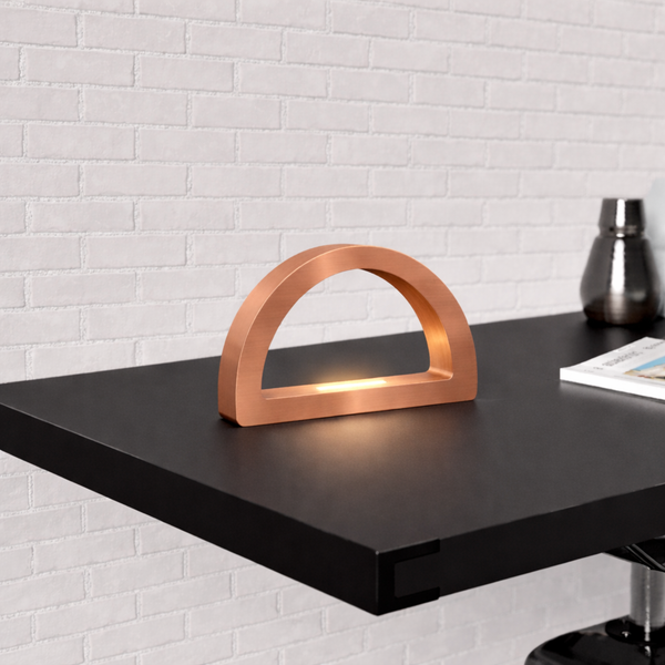 Copper finish arc-curved desk lamp by WOOM, casting a warm glow on a black desk with a white brick wall.
