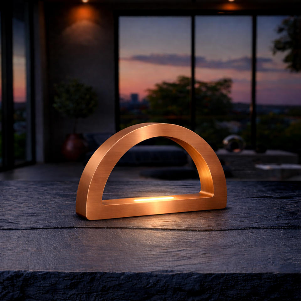 Copper finish arc-curved desk lamp by WOOM, glowing warmly on a dark textured table in a modern room.