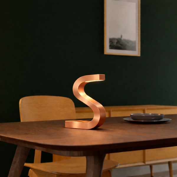 S-shaped copper finish table lamp by WOOM, casting a warm glow on a dark wood table.