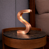 Copper finish S-shaped lamp by WOOM, standing on a round dark marble and oak wood side table.
