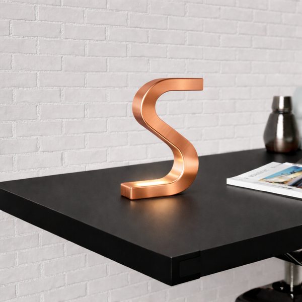 Copper finish S-shaped table lamp by WOOM, glowing at the base on a black shelf with a magazine.