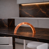 Arc-curved copper finish lamp by WOOM, glowing on a dark marble counter with orange veins.