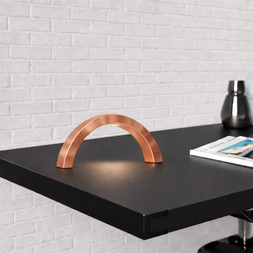 Arc-curved copper finish lamp by WOOM on a dark surface, casting a warm glow against a white brick wall.