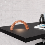 Arc-curved copper finish lamp by WOOM on a dark surface, casting a warm glow against a white brick wall.