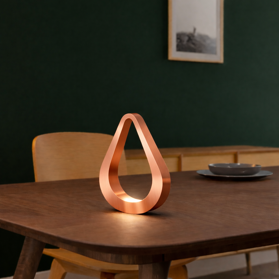 Copper finish handcrafted oak teardrop lamp by WOOM, glowing on a dark wooden table in a room with a green wall.