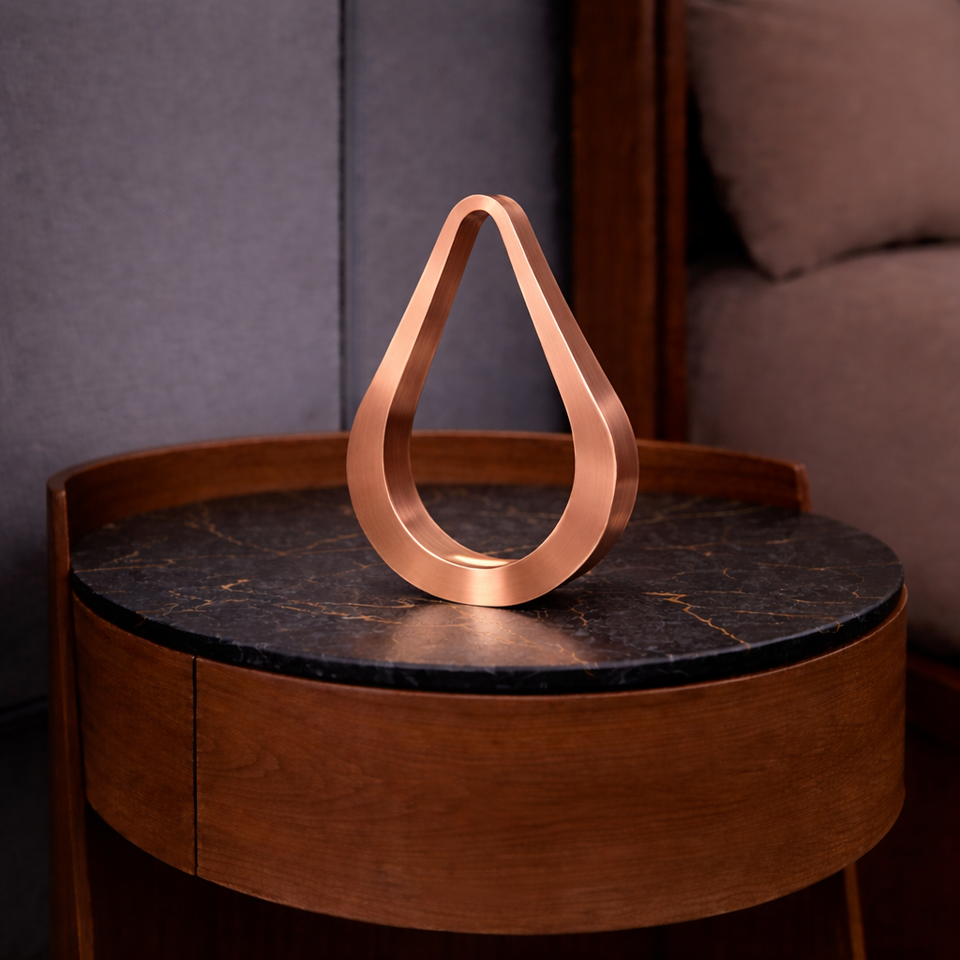 Copper finish teardrop shaped table lamp by WOOM on a dark marble-top wooden side table.