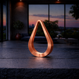 Teardrop shaped wooden lamp with a copper finish by WOOM, on a dark textured table in a modern room with a sunset view.