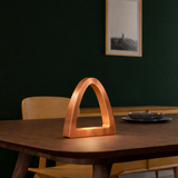 Copper finish handcrafted oak arch curved desk lamp by WOOM, glowing warmly on a dark wooden table.