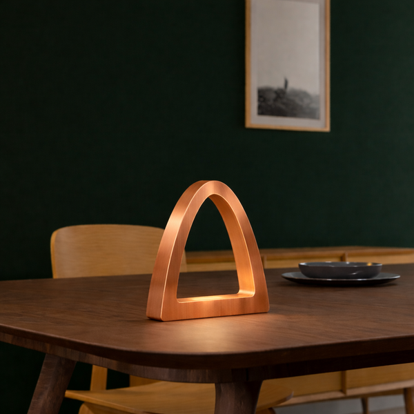 Copper finish handcrafted oak arch curved desk lamp by WOOM, glowing warmly on a dark wooden table.