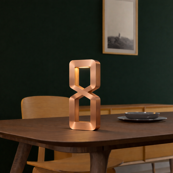 Copper finish hourglass-shaped table lamp by WOOM, casting warm light on a dark wooden table.