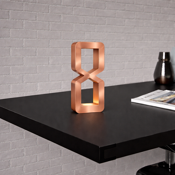 Copper finish hourglass-shaped table lamp by WOOM with integrated light, on a dark desk against a brick wall.