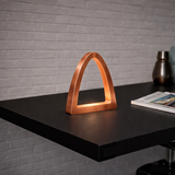 Copper finish arch curved desk lamp by WOOM, glowing warmly on a dark desk beside a magazine.
