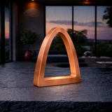 Copper finish arch curved desk lamp by WOOM, on a dark textured table in a modern room with a sunset view.
