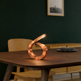 Copper-finished spiral desk lamp by WOOM with an illuminated tip, on a dark wooden table.
