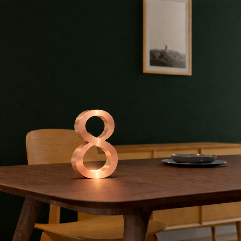 Copper finish infinity shaped table lamp from WOOM, emitting a warm glow on a dark wooden table with a dark green wall.