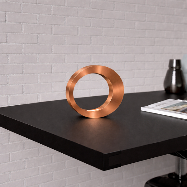Copper finish oval lamp by WOOM on a dark desk against a white brick wall, next to a magazine and dark bottle.