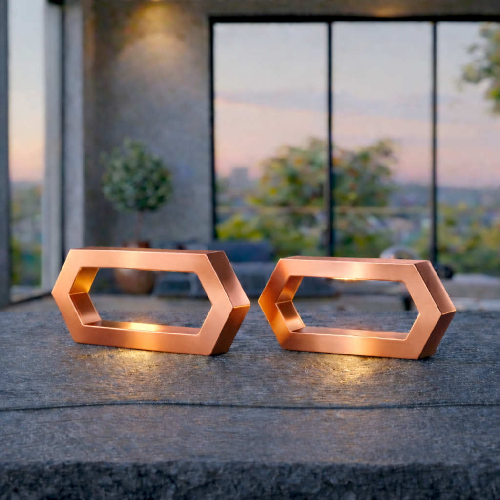 Two copper finish hexagon LED lamps by WOOM, glowing warmly on a dark stone table with a blurred outdoor patio at sunset.