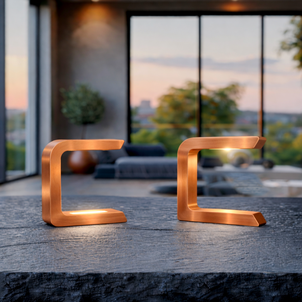  Two copper finish handcrafted wooden C-shaped lamps by WOOM, emitting warm light on a textured stone table.