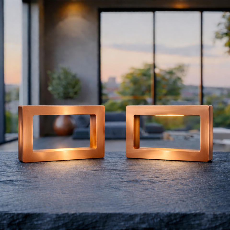  Two copper-finish rectangular LED lamps by WOOM, glowing warmly on a dark textured table.