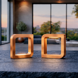 Two square copper finish wooden LED lamps by WOOM on a dark textured table in a modern room at sunset.