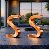 Copper finish S-shaped table lamp by WOOM, featuring two identical units casting a warm glow on a dark stone surface.