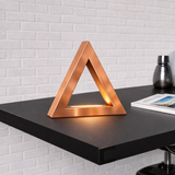 Copper finish handcrafted oak triangular lamp by WOOM with a warm glow on a black desk against a white brick wall.