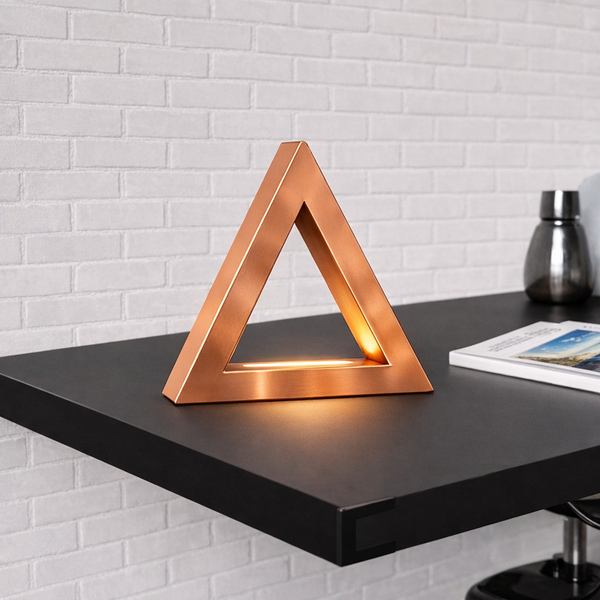 Copper finish handcrafted oak triangular lamp by WOOM with a warm glow on a black desk against a white brick wall.