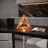 Copper finish triangular lamp crafted from oak by WOOM, with a warm internal glow on a dark marble countertop, next to an open cookbook.