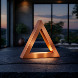 Copper finish triangular lamp by WOOM, glowing softly on a dark textured wooden table with an evening background.