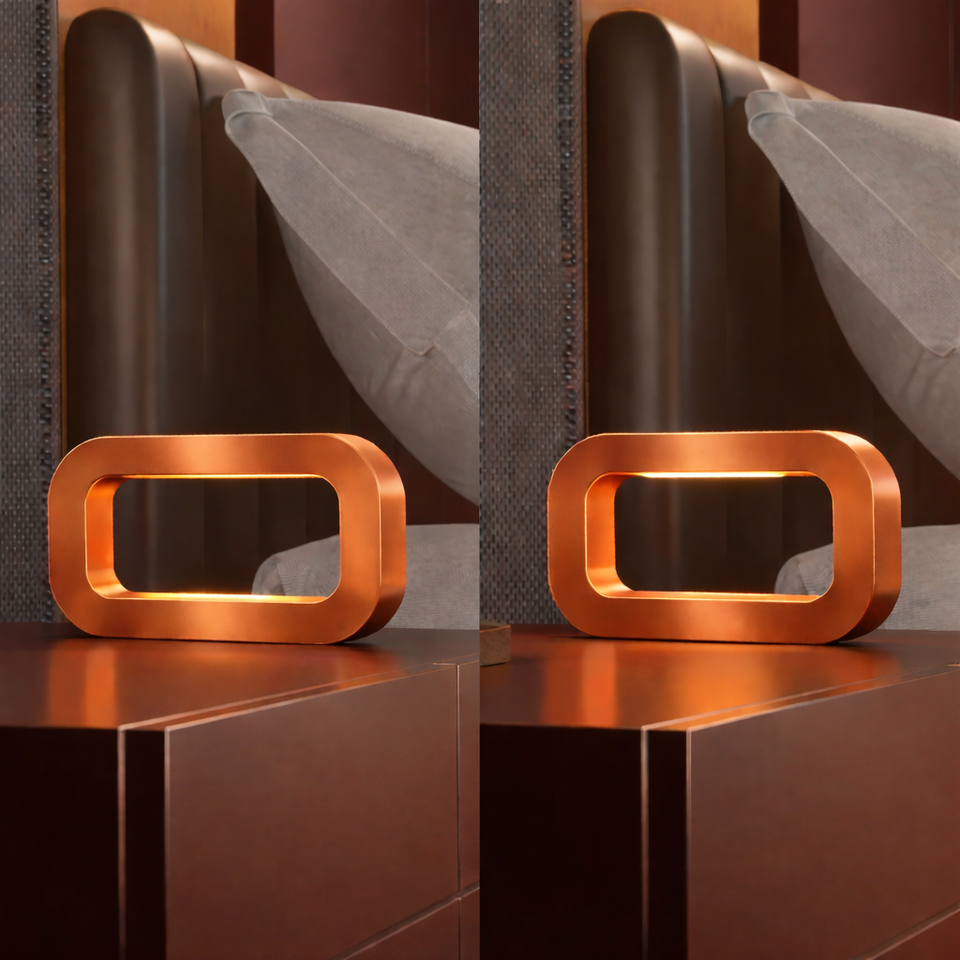 Copper finish rectangular table lamp by WOOM, with rounded corners and an illuminated interior on a dark wooden nightstand.